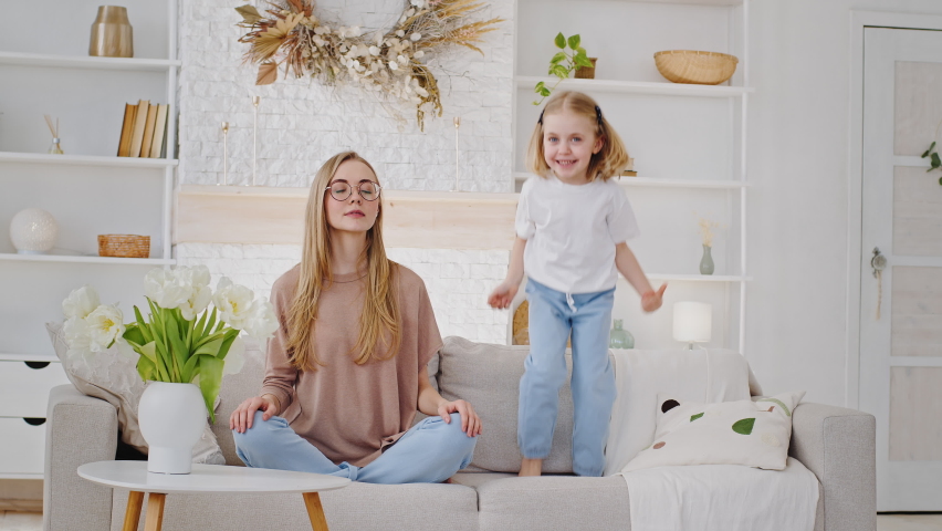 Noisy child active little girl daughter kid jumping on sofa baby bad behavior distracting disturbing young mother calm woman mom meditate sitting in lotus position in living room, tranquility concept Royalty-Free Stock Footage #1070215969