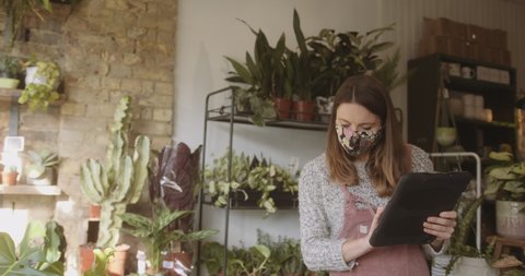 Young adult female small business sales clerk working on digital tablet in florist store wearing protective face mask