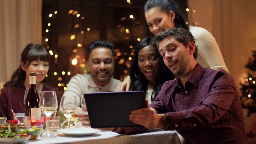 winter holidays, celebration and people concept - happy friends with tablet pc computer having video call at home christmas dinner