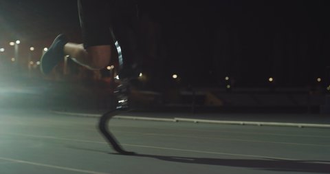 Cinematic close up of disable man with legs prosthesis running with effort with racing auto behind on car track at night. Concept of handicapped people active lifestyle, determination, motivation.