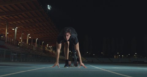 Cinematic shot of young disabled man with legs prosthesis prepares for launching off the start line on race track at night. Concept of handicapped people active lifestyle, determination.