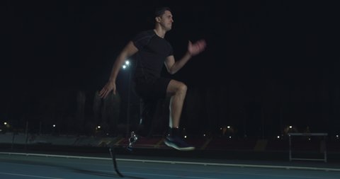 Cinematic shot of young disable man with legs prosthesis running with effort with racing auto behind on car track at night. Concept of handicapped people active lifestyle, determination, motivation.