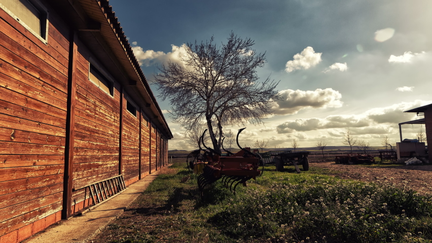 Timelapse of a sunset with clouds passing over a stable of horses. Tree and tools of field work