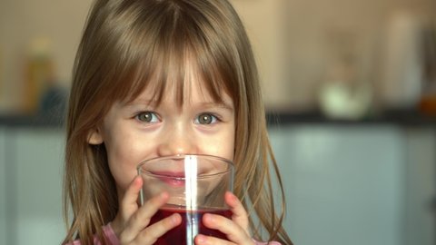Portrait of little girl, looks at camera, laughs and drinks cherry juice. Close up of happy child drinks fruit juice on kitchen background