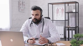 Male doctor wearing white coat consulting patient remote online using headset and web camera on laptop.Video call, video chat,conference with colleagues.Telemedicine.Remote healthcare services concept