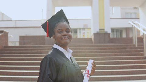 Young african american female graduate standing in front of the camera with a diploma and books in her hands. The student is dressed in a black robe and a square master's hat and stands outside.