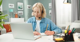 Portrait of Caucasian young pretty blonde woman in headset talking on webcam call on laptop studying online writing down information, female student having video lesson, webinar training concept