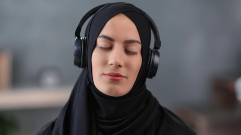 Closeup face of cheerful modern Islamic young woman enjoying audio sound wearing headphones having positive emotion. Joyful Muslim female in black hijab listening music. Shot with RED camera in 4K