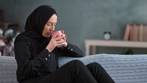 Satisfied Muslim young lady in traditional black hijab relaxing with hot mug of tea on couch. Beautiful Saudi female enjoy weekend at home having positive emotion. Shot on RED Raven 4k Cinema Camera