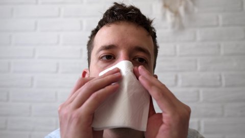 Sick guy is sitting on the bed and he has a runny nose. A man has the flu, a virus, or an allergy. Healthy medicine and people concept. Allergies, runny nose and sneezing. Blowing nose.