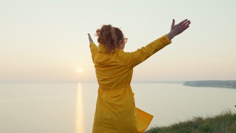 Happy Young Woman face Rejoices in Spring Weather Stands on Edge of Cliff on Seashore at Sunset Raises her Hands slow motion in Sky in Yellow Raincoat. Lifestyle freedom. Health