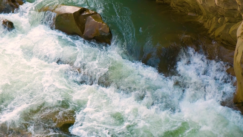 Aerial view of river waterfall with clear turquoise water falling down between wet boulders with thick white foam.