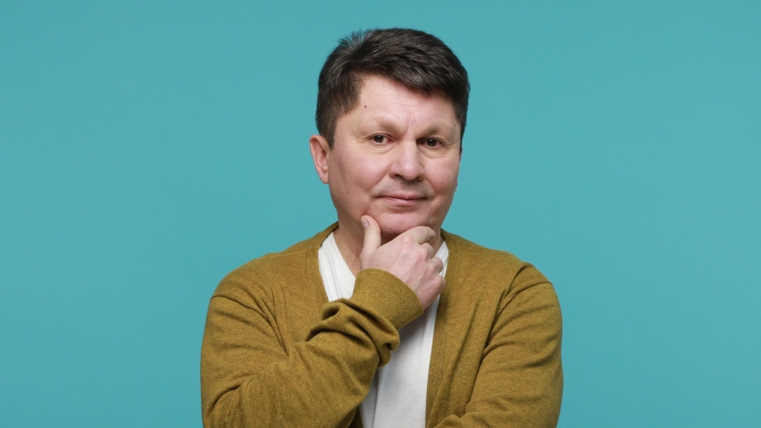 Hmm, need to think. Doubtful bewildered middle aged man in white t-shirt and cardigan contemplating rubbing his chin, hesitating making choice. Indoor studio shot isolated on blue background Royalty-Free Stock Footage #1070258800