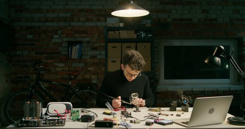 Focused Working Man is Soldering Electronic Circuit in Loft Workshop, IT Startup Office, Student Laboratory or Garage at Night. Technology Engineering for VR or DIY Concept. 4K Wide Pan Shot