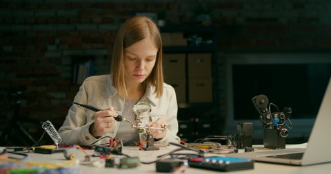 Focused IT Working Lady is Soldering Electronic Circuit in Loft Workshop, Startup Office, Student Laboratory or Garage at Night. Technology Engineering for VR or DIY Concept. 4K Medium Pan Shot