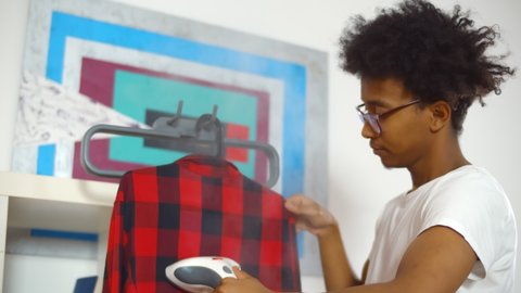 Young african man using steamer on checkered shirt before getting dressed. Afro-american guy in glasses steaming clothes on rack at home.
