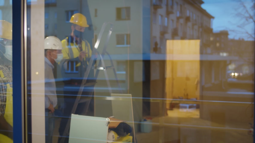 View through window of builders in safety mask bumping elbows greeting. African and caucasian construction workers in uniform and protective mask doing elbow bump at construction site Royalty-Free Stock Footage #1070268202