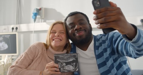 Happy multiracial married couple holding ultrasound picture of baby and taking selfie. Pregnant caucasian woman and african man taking selfie on smartphone with sonogram image
