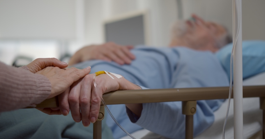 Sick senior man in hospital bed and his wife sitting next to him. Close up of aged woman holding hand of ill husband with intravenous drip lying in hospital ward Royalty-Free Stock Footage #1070268358