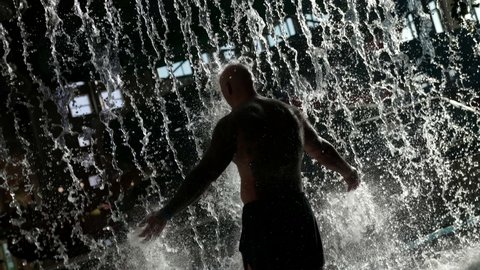 In the water park behind the waterfall there is a big bald man, doused with water, he is in swimming trunks, a view from the back.