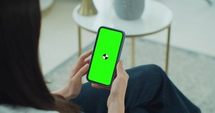 Female using phone while sitting on couch at home. Woman holding and looking at smartphone mock up screen. Concept of chroma key and green screen.