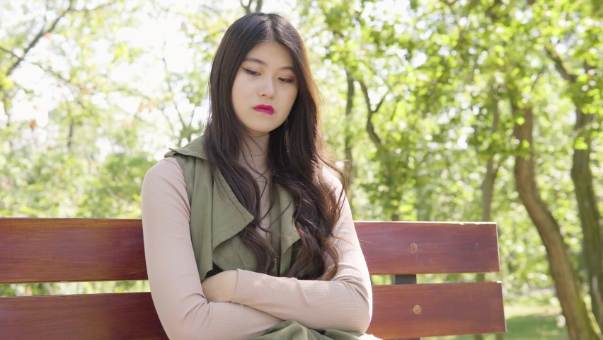 A young Asian woman thinks about something as she sits on a bench in a park on a sunny day | Shutterstock HD Video #1070281591