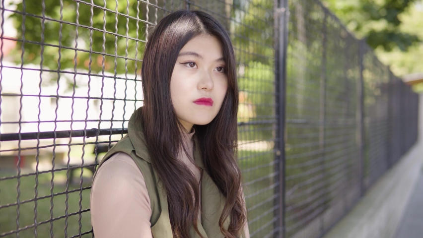 A young Asian woman thinks about something and waits for someone as she leans against a tall fence in an urban area - closeup | Shutterstock HD Video #1070281648