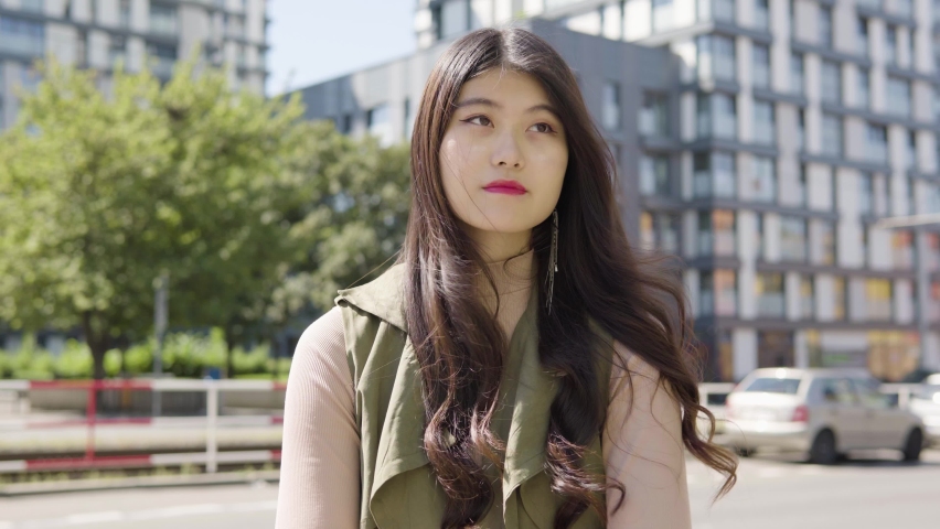A young Asian woman thinks about something as she waits in a street in an urban area - cars drive on a busy road in the blurry background | Shutterstock HD Video #1070281702