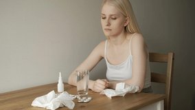 Medium shot of blond woman with sore throat sitting at kitchen table and coughing.