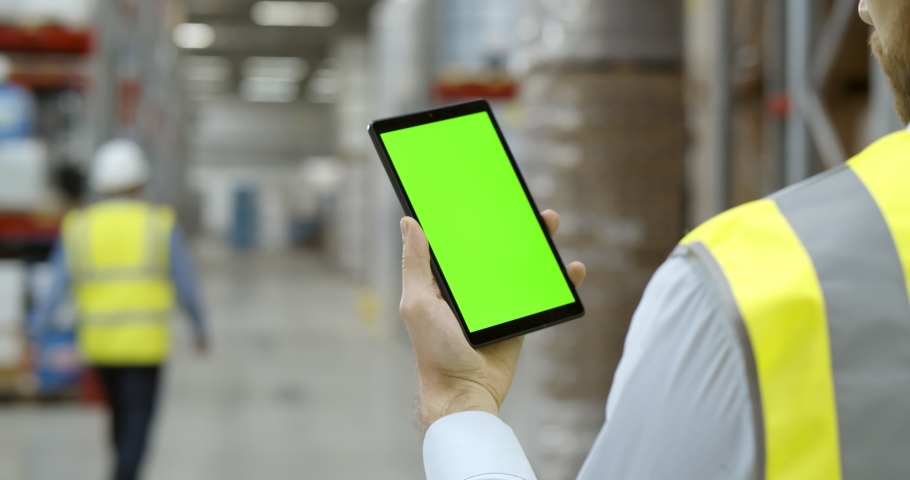 Back view of male worker in warehouse wearing safety vest standing near shelf with goods using cellphone. Cropped shot of factory storehouse manager using mobile phone. Green screen Royalty-Free Stock Footage #1070283766
