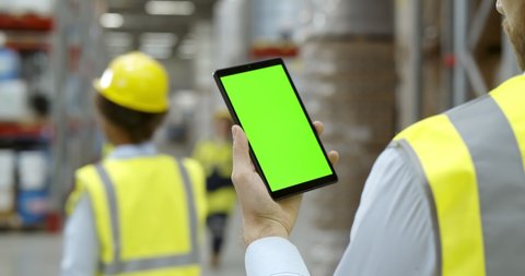 Back view of male worker in warehouse wearing safety vest standing near shelf with goods using cellphone. Cropped shot of factory storehouse manager using mobile phone. Green screen