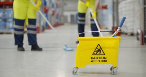 Cropped shot of worker swearing safety overall holding mop cleaning floor in warehouse. Team of janitors with bucket washing floor in industrial storehouse