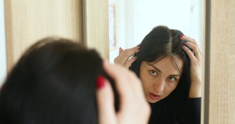 Portrait of a beautiful young woman examining her scalp and hair in mirror, hair roots, color, first grey hair, hair loss or dry scalp problem, or noticing that she is suffering from dandruff