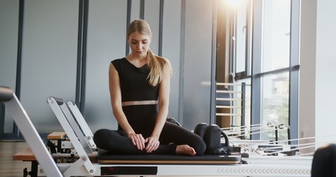 Fit athletic young woman sitting on a pilates reformer machine in a gym tying her long blond hair backlit by a glowing sun through a large window in an active lifestyle or health and fitness concept
