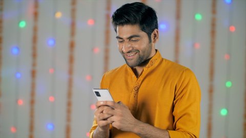 Attractive man in traditional Indian wear smiling and texting on a smartphone - Festive decor. Bokeh shot of a young Asian happy guy wearing a yellow kurta chatting over the phone and smiling - Diw...