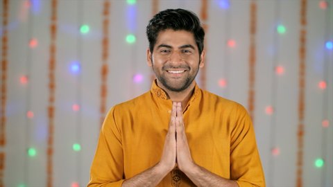 Young man greeting in traditional Indian namaste while looking at the camera. Bokeh shot - Handsome Indian guy happily joining hands in namaste gesture on Diwali festival celebrated in India