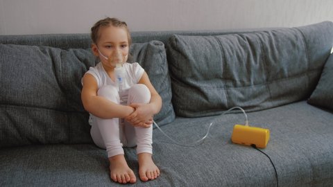 Little girl makes inhalation with medical nebulizer while sitting on bed. Cute child is sick bronchitis or asthma and breathes through inhaler at home