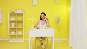 Young Caucasian lady, happy office worker, in white top is sitting at workplace in yellow office, having pleasant phone talk, discussing last news with friend, uses landline telephone, Slow motion.
