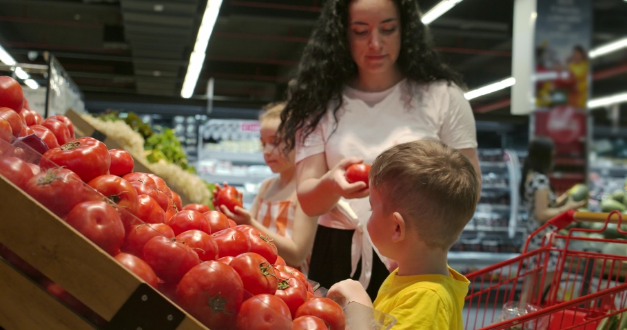 Cute woman with little happy son and daughter are choosing fresh vegetables together at the grocery store. A mother with her children takes ripe tomatoes from the shelf and puts them in a basket. Royalty-Free Stock Footage #1070307574
