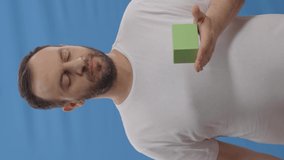 Young man with a beard in front of a blue background is showing the green little box in his left hand with his right index finger and looking at the screen and laughing.Video for the vertical story.