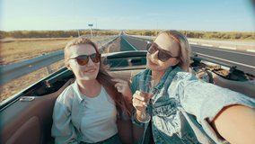 Beautiful woman takes video on her phone as she and her friend ride in convertible and drink champagne in back seat. Girl takes selfie while riding with girlfriend in convertible with an open roof
