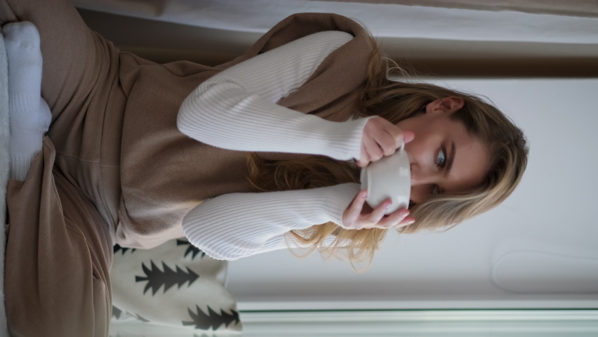 A young beautiful blonde caucasian woman relaxes sitting on the windowsill in home. A woman drinks tea or coffee from a white cup in a cozy interior. The concept of a comfortable serene and good morni