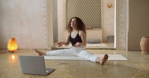 Young Curly-Haired woman yoga instructor coaching online, making video of yoga lessons on laptop camera. Attractive fit girl watching yoga lesson in laptop.