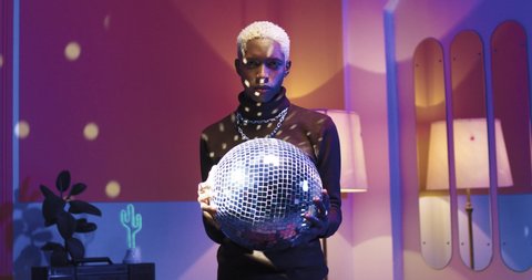 Portrait of handsome African American young male standing in room in evening at home holding big disco ball in hands looking at camera, disco lighting, vintage style, leisure, party time concept