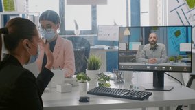 Freelancer wearing protective face mask during online videocall sitting at desk in startup company office. Coworkers working on computer during coronavirus global pandemic