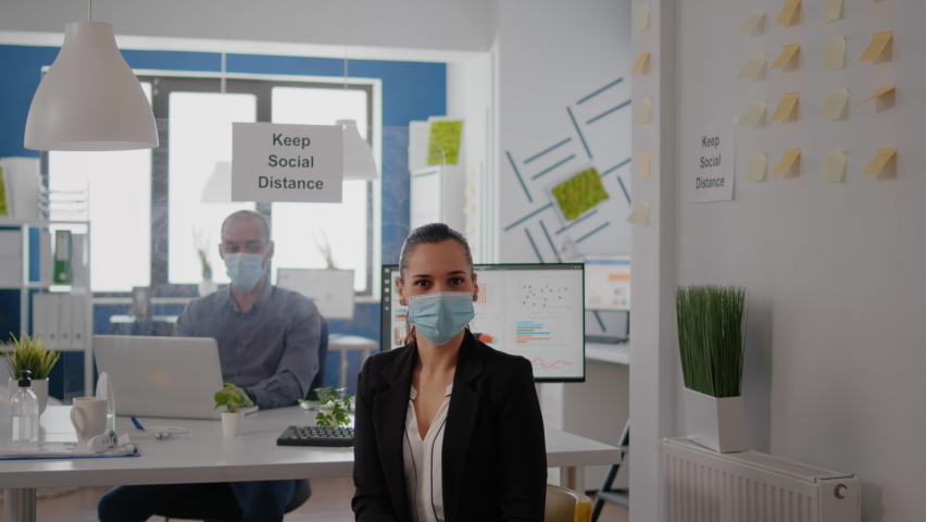 Portrait of woman manager wearing face mask to prevent infection with coronavirus sitting on chair at desk table in business ofice. Colleagues maintain social distancing using separeted plastic board