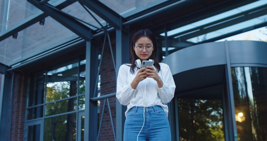 Young dark haired woman with headphones standing in business buildings district. Pretty Asian female texting, messaging and browsing on smartphone in city street. Technology concept.