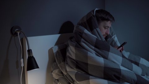 Young man, cell phone addict, wakes up late at night in bed wrapped upside down in blanket using smartphone and cannot sleep, suffers from insomnia. Gadget, social network addiction. Sleep disorder