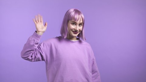 Friendly woman waving hand - hello. Greeting, say Hi to camera. Beautiful young girl with dyed purple hair on violet studio background.