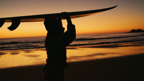 Handheld footage of a woman carries a surfboard on a sunset beach after sundown. Beautiful and cinematic shot of surfing wanderlust lifestyle. Vídeo Stock